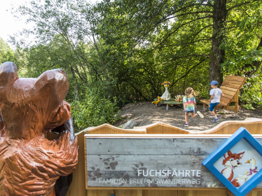 Schild Erlebniswanderweg Fuchsfährte mit spielenden Kindern im Hintergrund